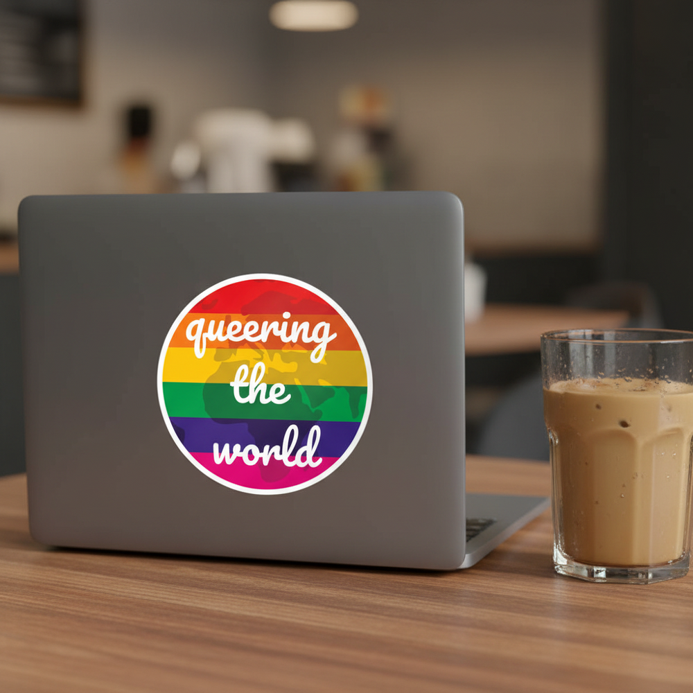 Queering The World Bubble-Free Sticker in rainbow colors on a gray laptop, wooden table, iced coffee beside it