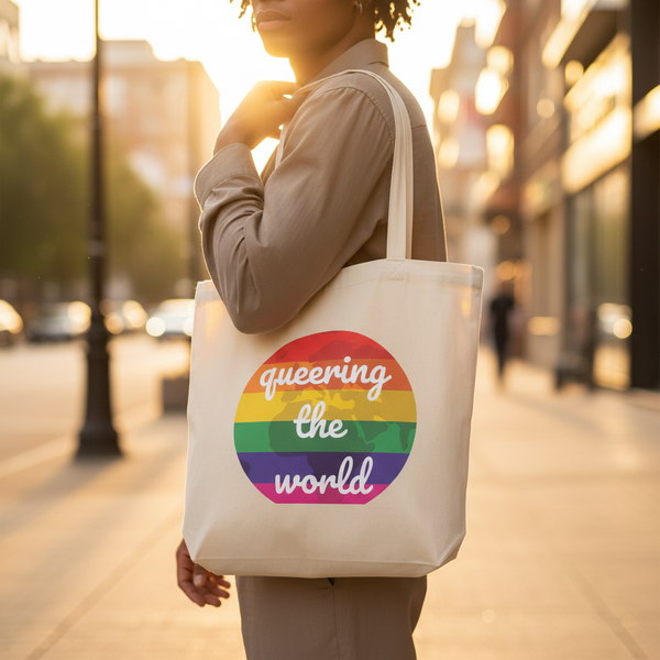 Eco tote bag with a rainbow circle and “queering the world” in white, held outside on a sunny city street; queer pride vibes.