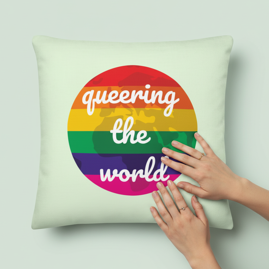 Light-colored pillow with a round rainbow world map, “queering the world” in white cursive, held by two ringed hands.