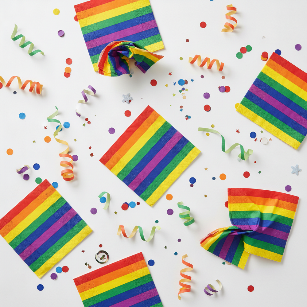 Overhead view of rainbow-striped paper napkins scattered with colorful party confetti and streamers