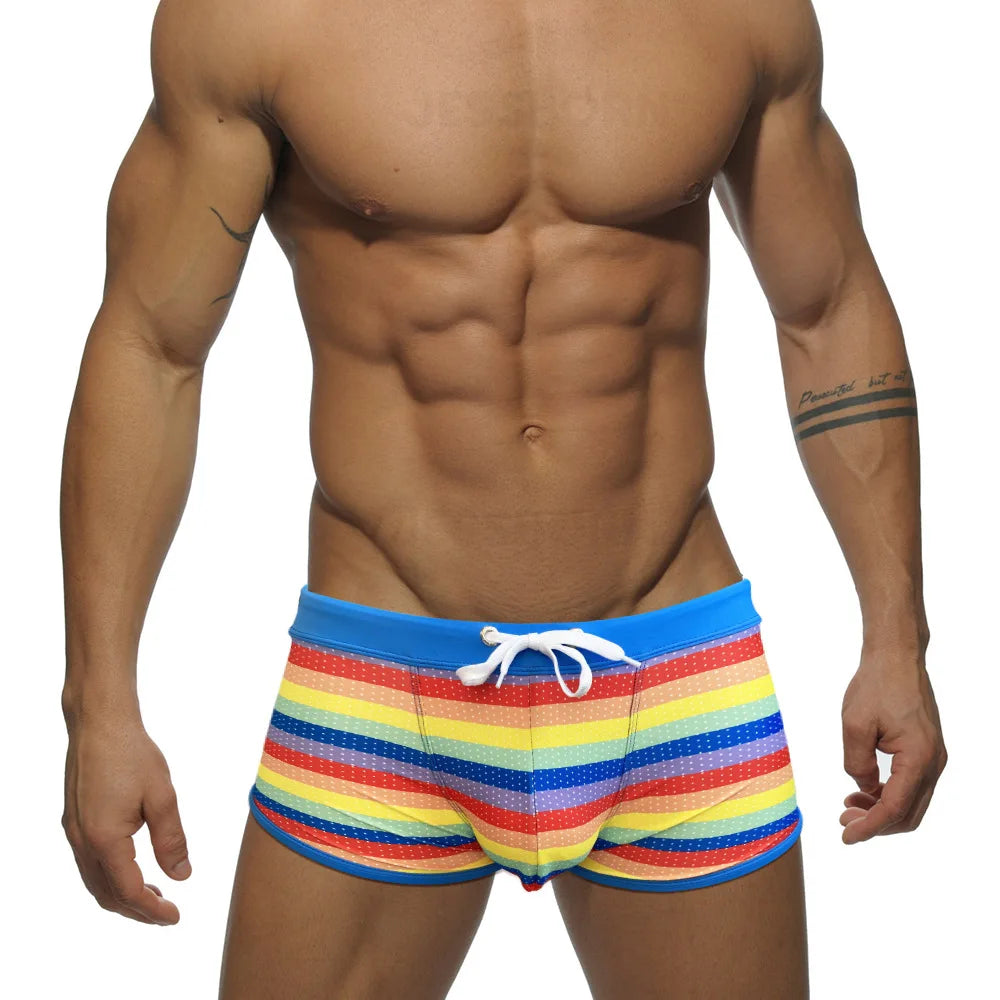 Front view of a model wearing rainbow-striped square-cut swim trunks with a white drawstring