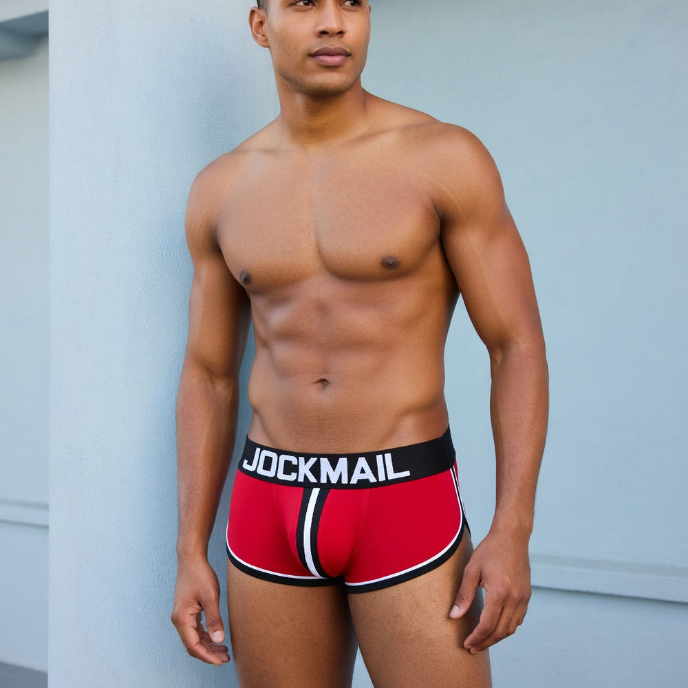 Adult model wearing red Jockmail sports boxers with black waistband and white piping against a pale blue wall.