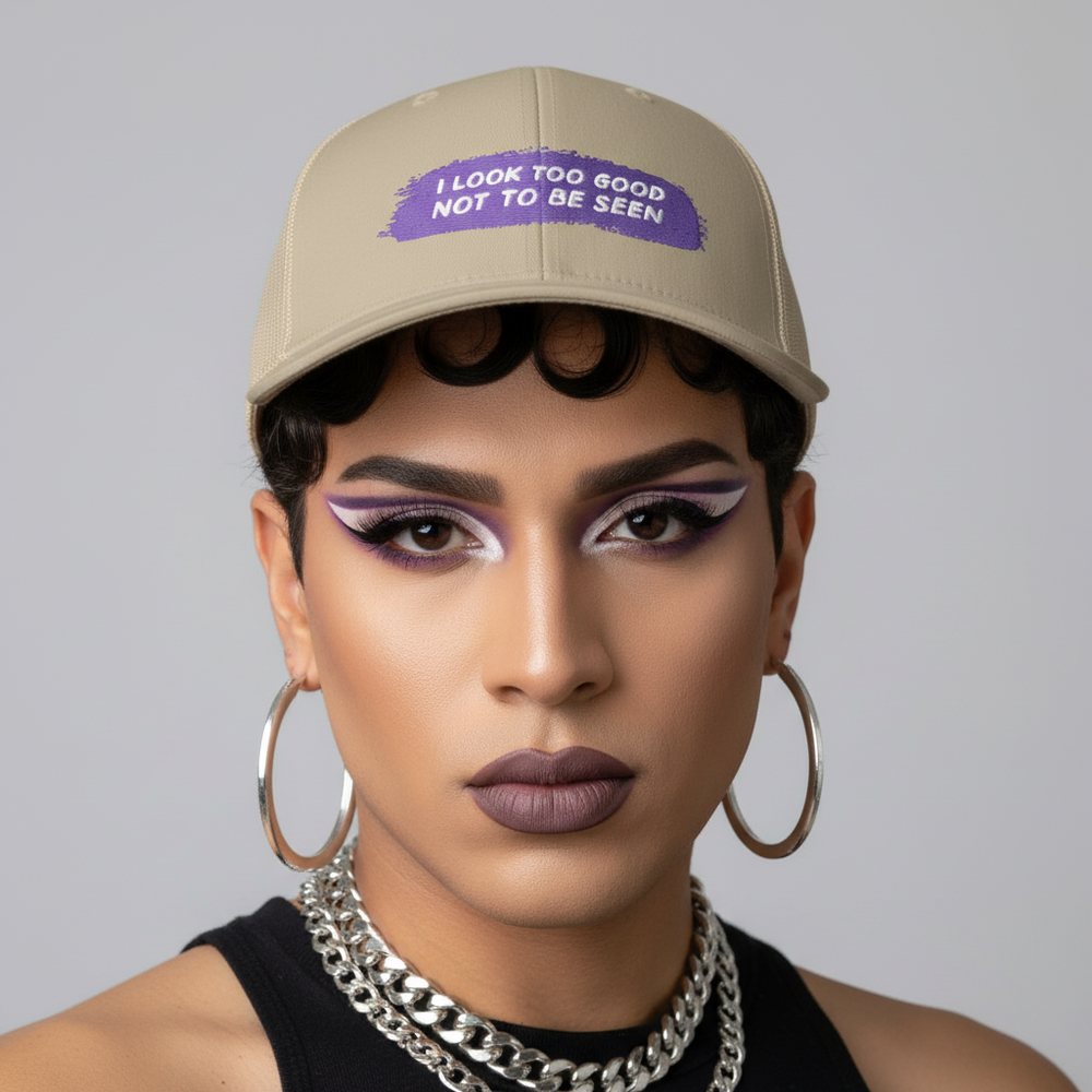 Beige trucker cap with “I Look Too Good Not To Be Seen” in purple, worn by someone serving bold makeup and queer style.