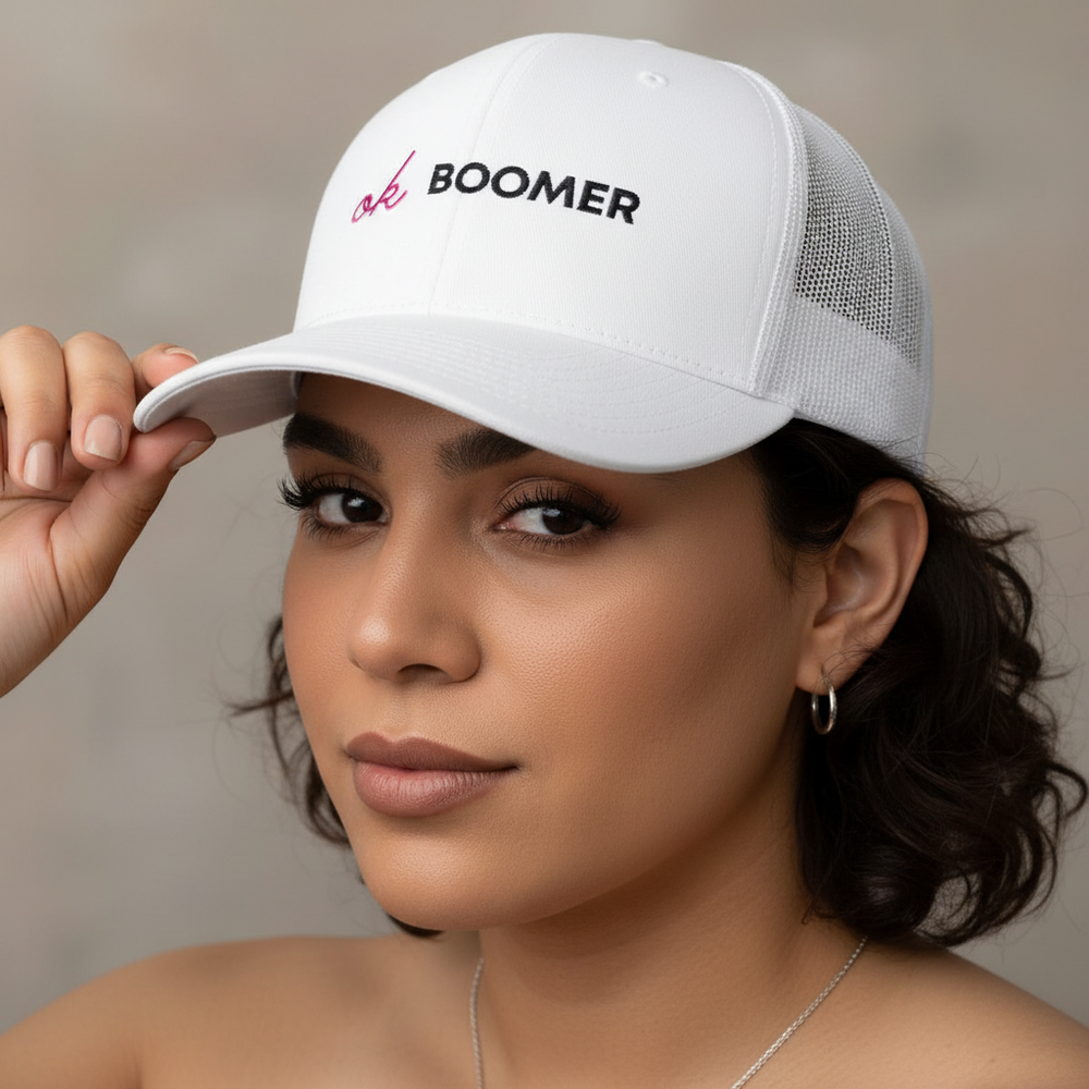 Trucker hat with white mesh back and “ok BOOMER” in black and pink worn by a femme with short dark hair, confidently posing.