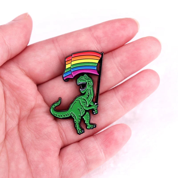 Hand holding a T. rex enamel pin with a rainbow Pride flag
