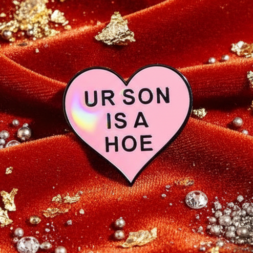 Heart-shaped pink enamel pin reading “UR SON IS A HOE” on red velvet