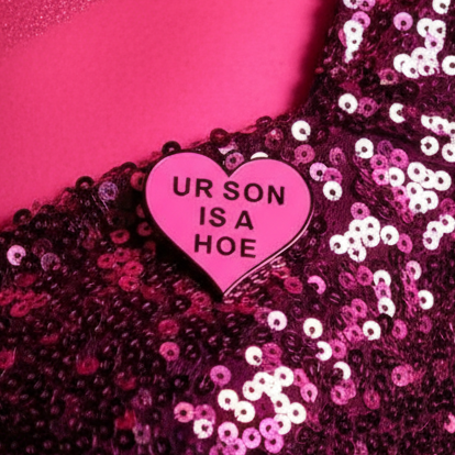 Pink heart enamel pin reading “UR SON IS A HOE” on magenta sequined fabric
