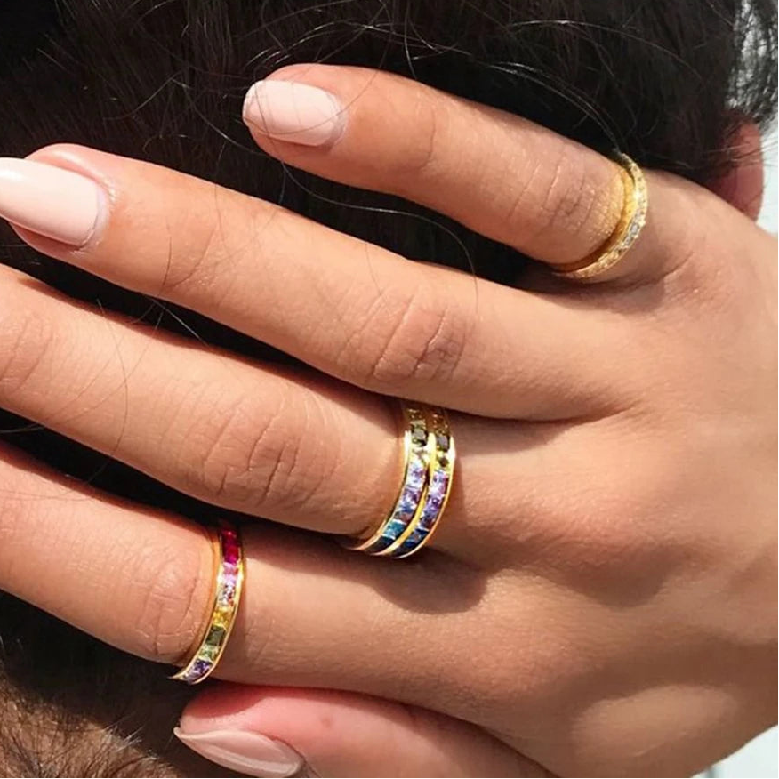 Hand wearing a rainbow-jeweled ring alongside two gold bands