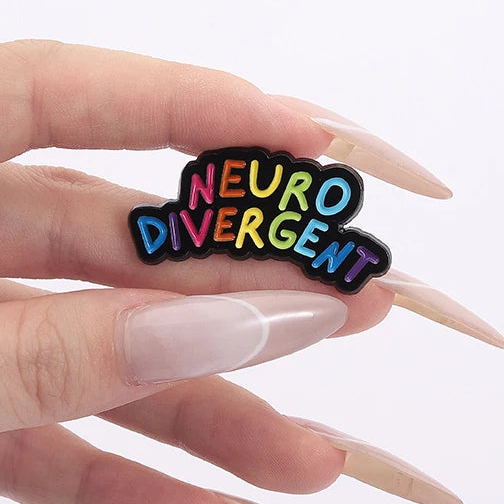Hand holding a black enamel pin reading “NEURODIVERGENT” in rainbow letters