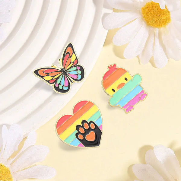 Three colorful enamel pins: butterfly, rainbow-striped duck, and rainbow heart with a paw print