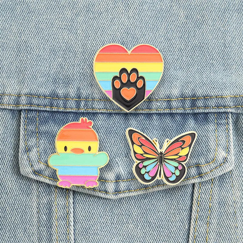 Three rainbow enamel pins—a heart paw, striped duck, and colorful butterfly—on a denim pocket