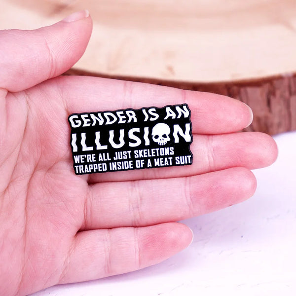 Enamel pin reading “GENDER IS AN ILLUSION” with a skull and skeleton slogan, held in a hand