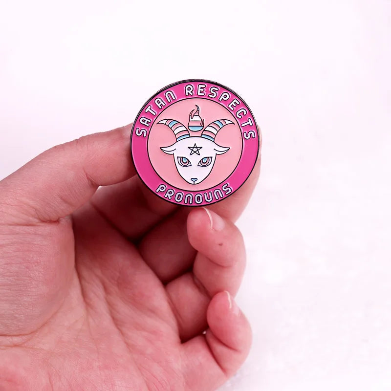 Hand holding a pink enamel pin reading “SATAN RESPECTS PRONOUNS,” with a horned goat design