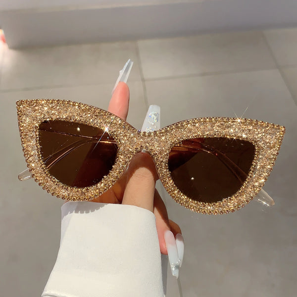A hand holds gold rhinestone cat-eye sunglasses with dark tinted lenses