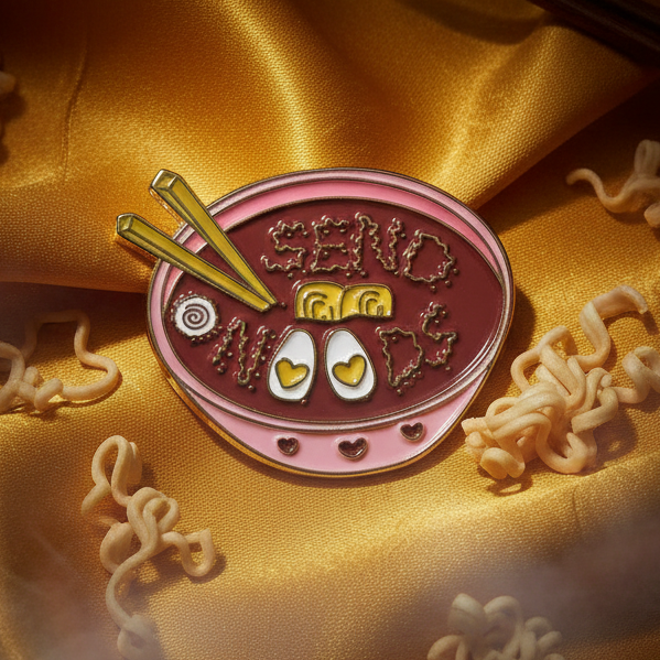 Pink oval enamel pin with yellow chopsticks, noodles, and a bowl-shaped design reading “SEND NOODS”