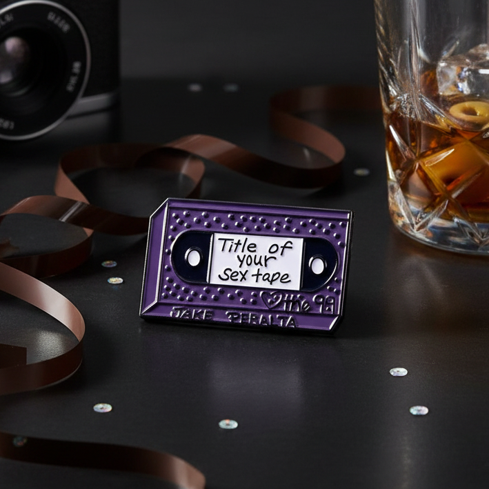 Purple cassette tape enamel pin reading “Title of your Sex Tape” on a dark tabletop