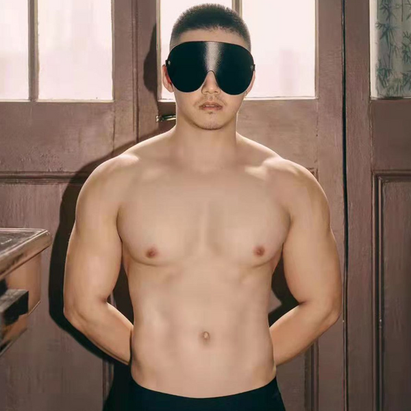 Shirtless model facing forward in a glossy black leather-like eye mask indoors