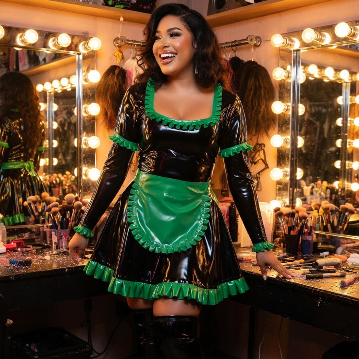 A person in a sexy French fetishist apron dress smiles at a bulb-lit makeup vanity