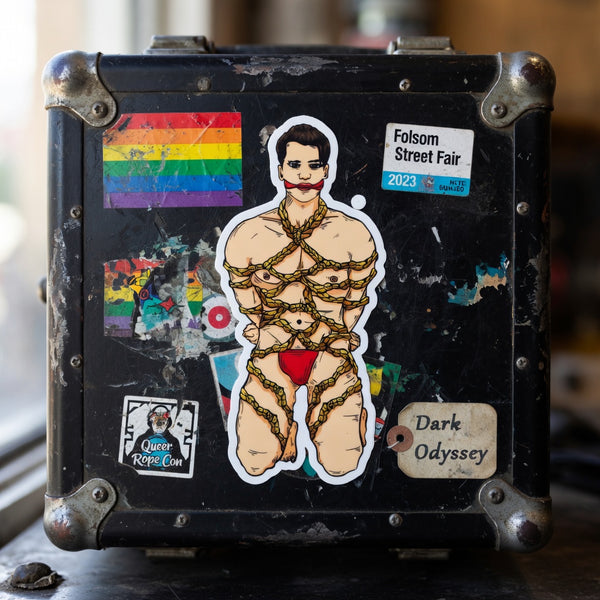 Close-up of a worn black suitcase with a central shibari-bound figure sticker and colorful stickers