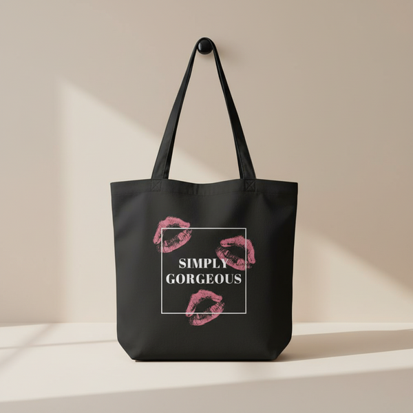 Black organic cotton tote bag with bold pink lipstick kisses and “SIMPLY GORGEOUS” in white, hanging on a wall.