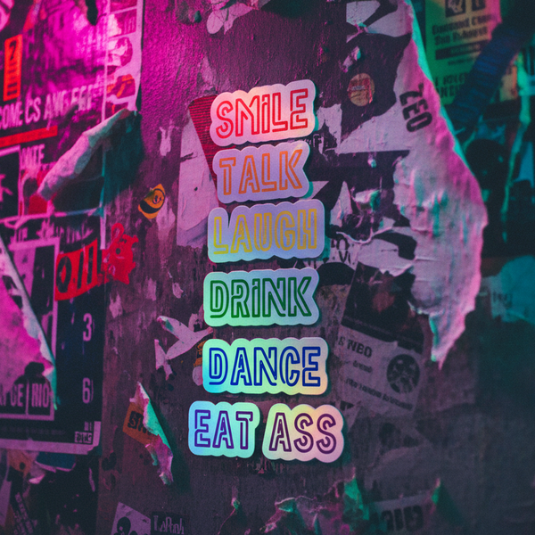 Rainbow holographic vinyl stickers reading “Smile, Talk, Laugh, Drink, Dance, Eat Ass” pop against a queer graffiti wall.