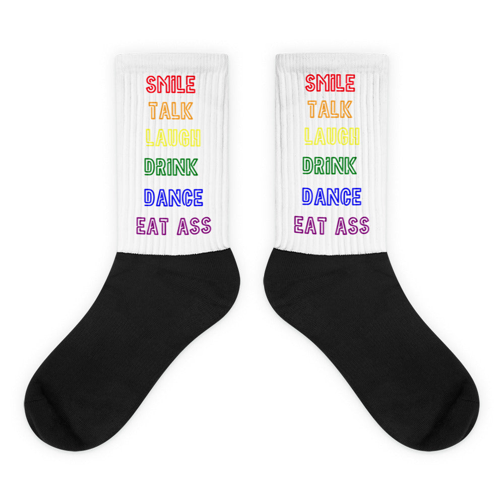 Pair of black-and-white crew socks with rainbow text reading “SMILE TALK LAUGH DRINK DANCE EAT ASS”
