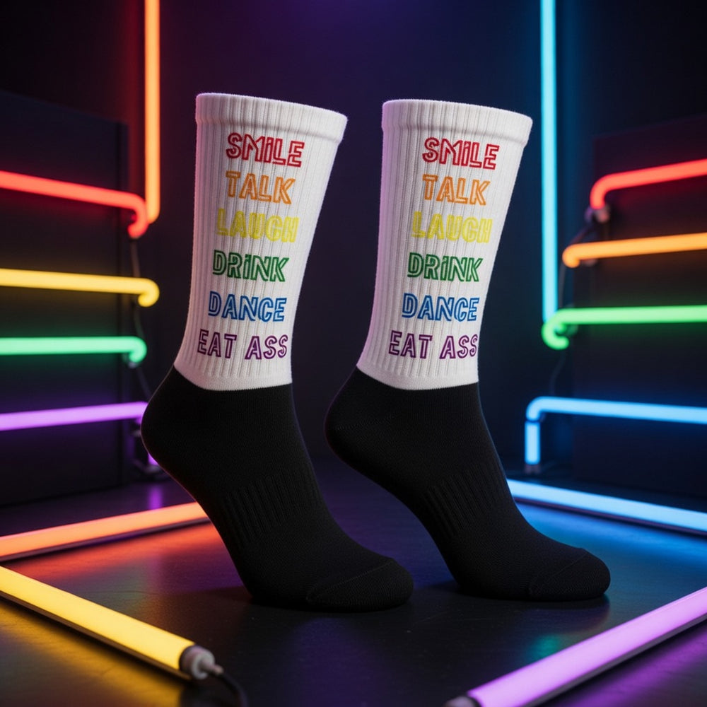 White and black crew socks read "Smile, Talk, Laugh, Drink, Dance, Eat Ass" in bold rainbow letters; neon tubes glow behind.