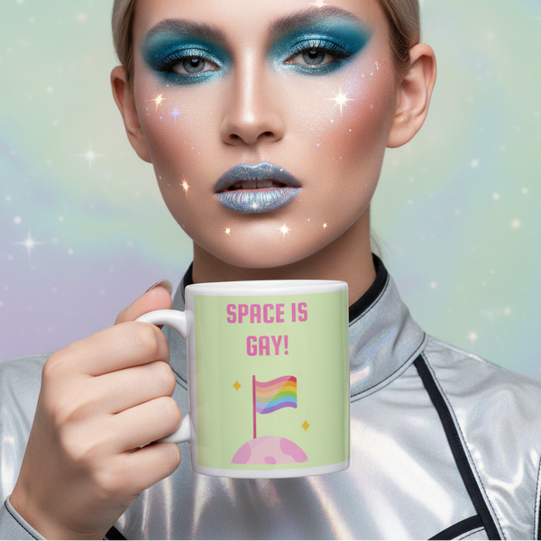 Person with cosmic makeup holds a mug reading “SPACE IS GAY!” with a rainbow flag