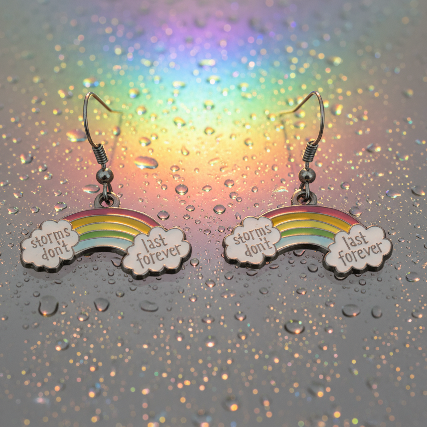 Rainbow earrings reading “storms don’t last forever,” photographed on a water-dappled glitter background