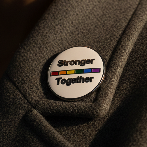 Close-up of a round enamel pin reading “Stronger Together” with a rainbow stripe