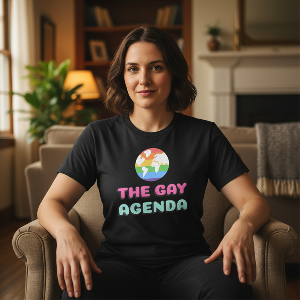 Smiling person seated indoors wearing a black T-shirt reading “THE GAY AGENDA”