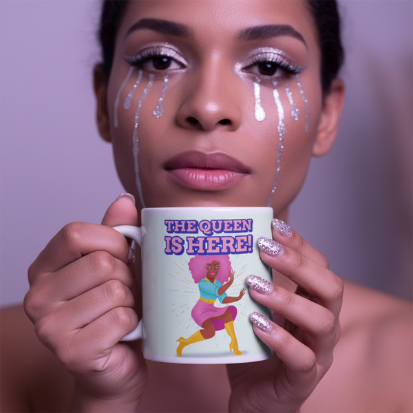 Close-up person holds a mug reading “THE QUEEN IS HERE!” with a dancing queen illustration