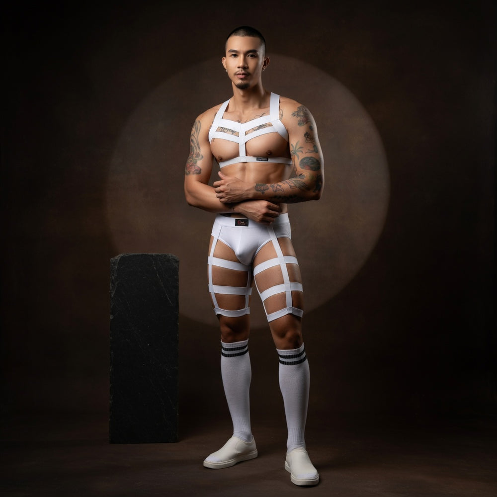 Male model wearing The Works white elastic chest harness, matching briefs, triple thigh straps and striped socks in a studio campaign.