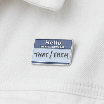 Blue enamel pronoun pin reading “Hello, my pronouns are They/Them” on white fabric
