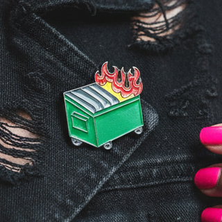 Close-up of a green dumpster-fire enamel pin on distressed black fabric