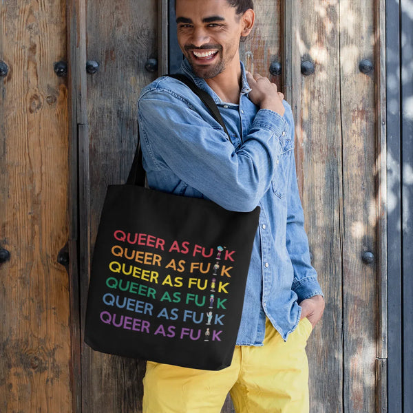 Model carrying a black tote bag printed with repeated rainbow “QUEER AS FU$K” lettering