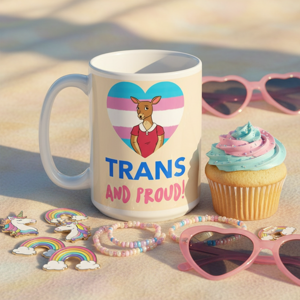 White mug reading “TRANS AND PROUD!” beside a pastel-frosted cupcake and pride-themed accessories