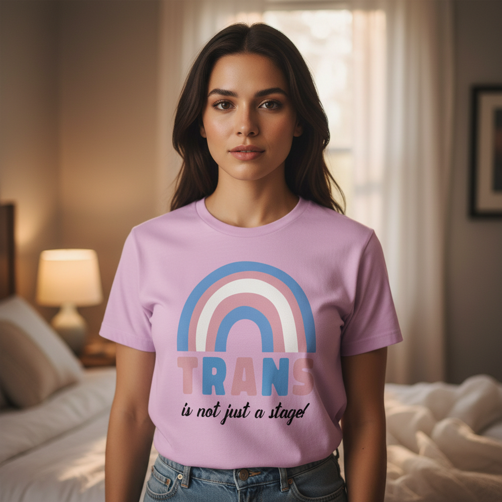 Lavender "Trans Is Not Just A Stage!" tee with trans pride rainbow, bold letters, worn by a person in a softly lit bedroom