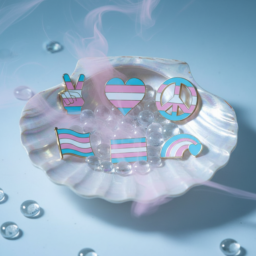 Six transgender pride enamel pins arranged in an iridescent shell dish