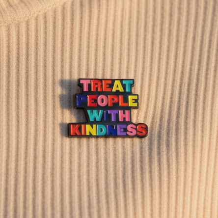 Rainbow enamel pin reading “TREAT PEOPLE WITH KINDNESS” on a wood-grain surface