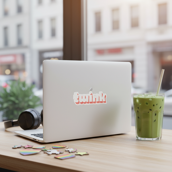White laptop displaying a rainbow-edged magnet reading “twink” on a café table
