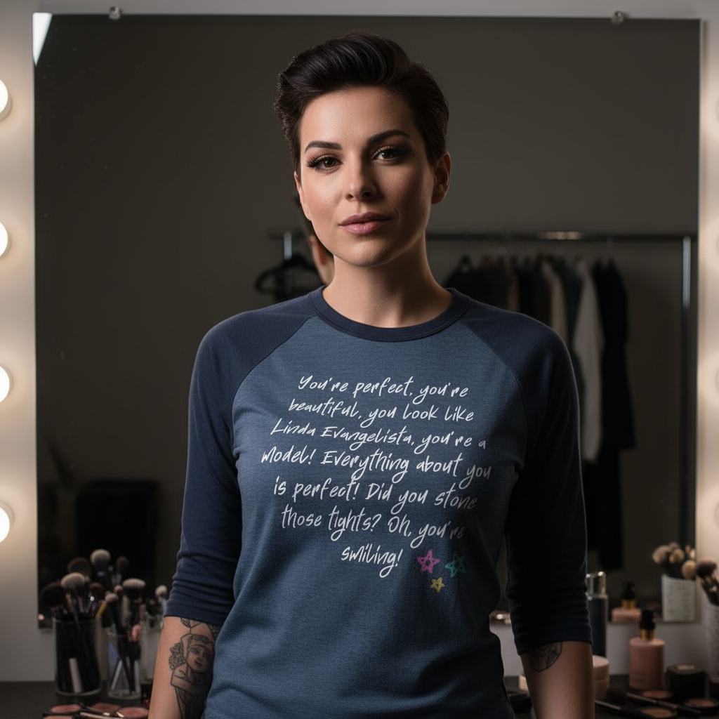 The You Look Like Linda Evangelista 3/4 Sleeve Raglan Shirt features bold script on a stylish blue raglan, celebrating queer pride and self-expression amidst a vibrant makeup station backdrop.