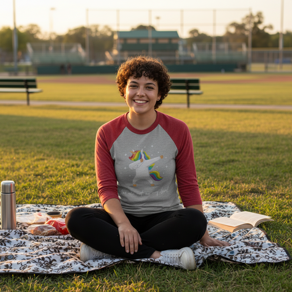 Smiling person seated on a picnic blanket wearing a gray raglan shirt with a rainbow-maned unicorn graphic