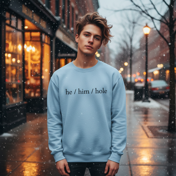 Young person wearing a light blue “he / him / hole” sweatshirt on a snowy city sidewalk