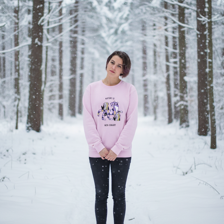 Person wearing a pink sweatshirt with a colorful creature and “Nature Is Non-Binary” text in a snowy forest