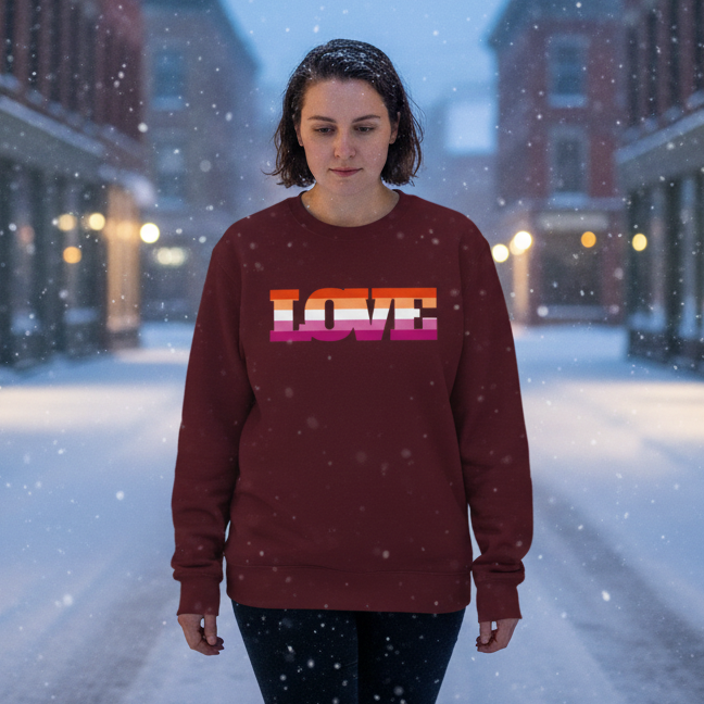 Young person in snow, wearing the Lesbian Love Unisex Sweatshirt in maroon with vibrant “LOVE” stripes. Showcases queer pride amidst softly lit storefronts, embodying warmth and self-expression in a cozy winter scene.