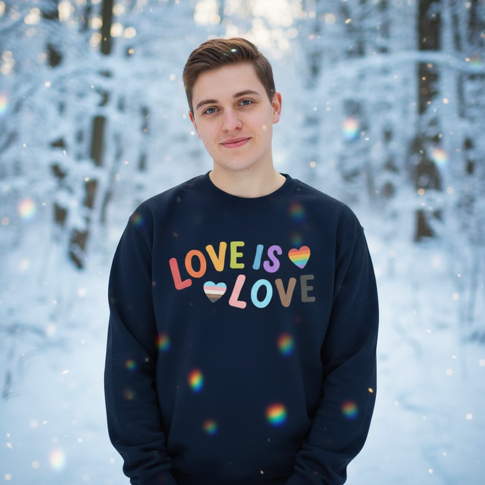 Person wearing a navy sweatshirt reading “LOVE IS LOVE” with rainbow hearts in a snowy forest