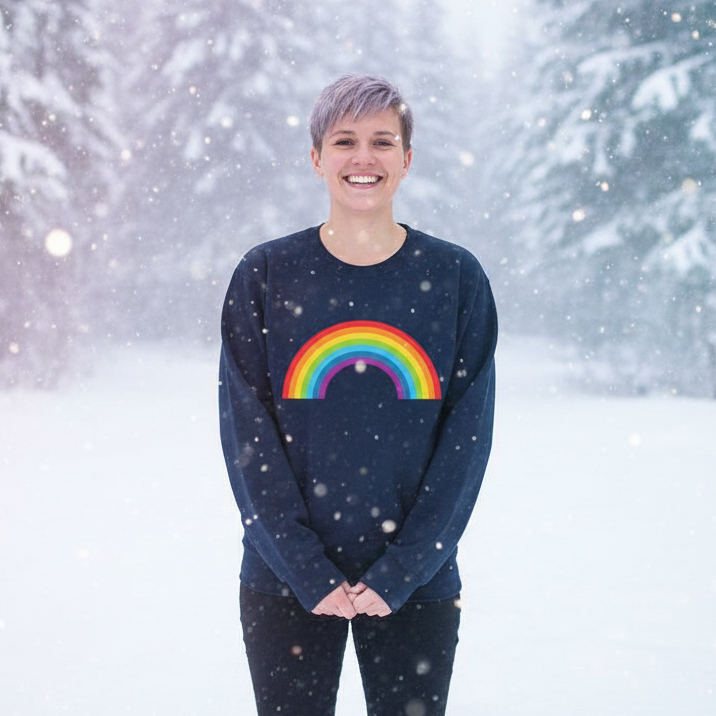 Person smiling outdoors in snowy forest, wearing Rainbow Unisex Sweatshirt with bold, arched rainbow graphic, embodying queer pride and self-expression. Ideal for warmth, comfort, and visibility in colder months.
