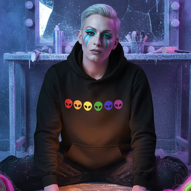 A person with platinum-blonde hair wears the Gay Alien Unisex Hoodie—black with rainbow alien-head icons—exuding queer pride and playful style, reflected in a frosted vanity setting with bold makeup.