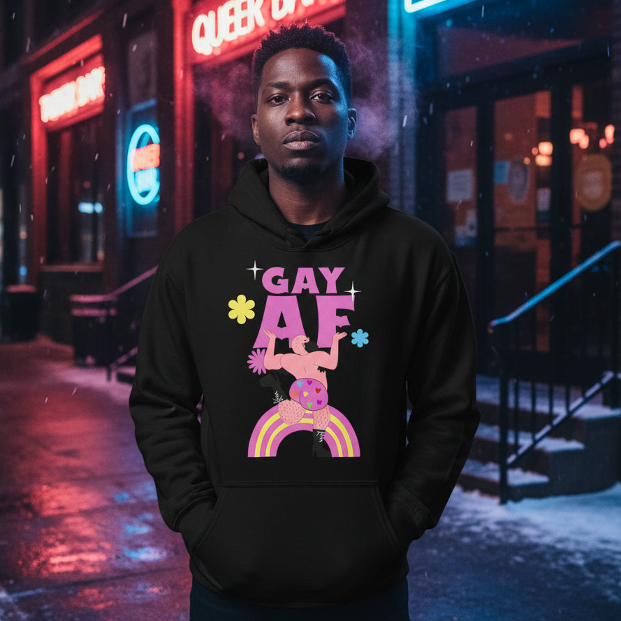 Young man wearing the Gay AF Unisex Hoodie, featuring a vibrant graphic with a muscular figure and rainbow accents, standing confidently on a neon-lit city street at night, reflecting pride and self-expression.
